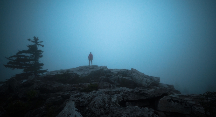 A person stands on a rocky overlook in dense fog. 